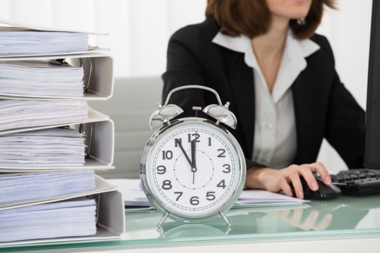 Close-up Of Young Businesswoman Working On Computer In Office With Alarm Clock On Desk