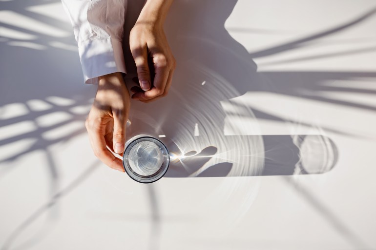 Woman's hands and a glass of clean water on the white table in natural sunlight with plant shadows. Minimalist lifestyle concept