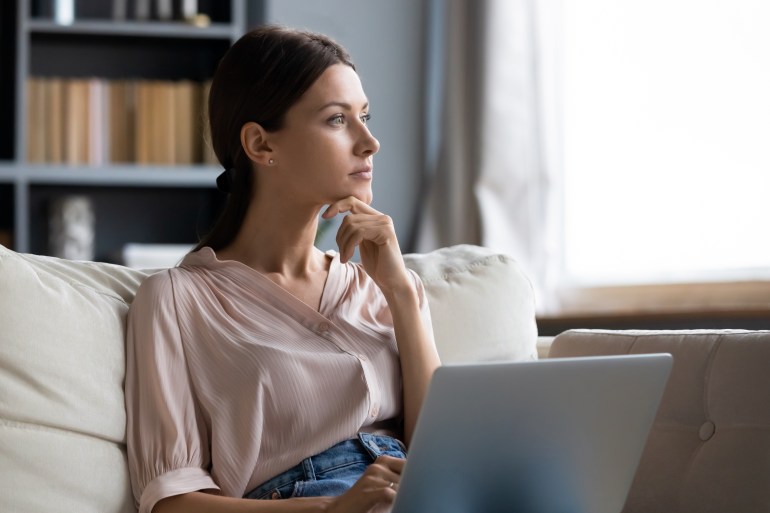 Distracted from work worried young woman sitting on couch with laptop, thinking of problems. Pensive unmotivated lady looking at window, feeling lack of energy, doing remote freelance tasks at home.