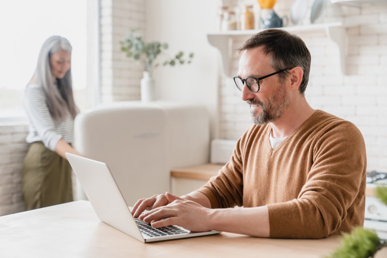 Mature husband man freelancer businessman working from home on laptp typing searching web, e-learning while his middle-aged wife doing household chores in the kitchen on lockdown; Shutterstock ID 1998725270; purchase_order: aljazeera ; job: ; client: ; other: