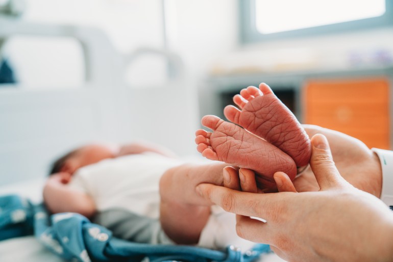 A mother is holding the feet of her newborn baby at the hospital. She's sitting on the bed at the hospital.